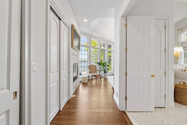 A hallway with white doors, a wooden floor, a chair, a plant, and a window with a view of greenery.