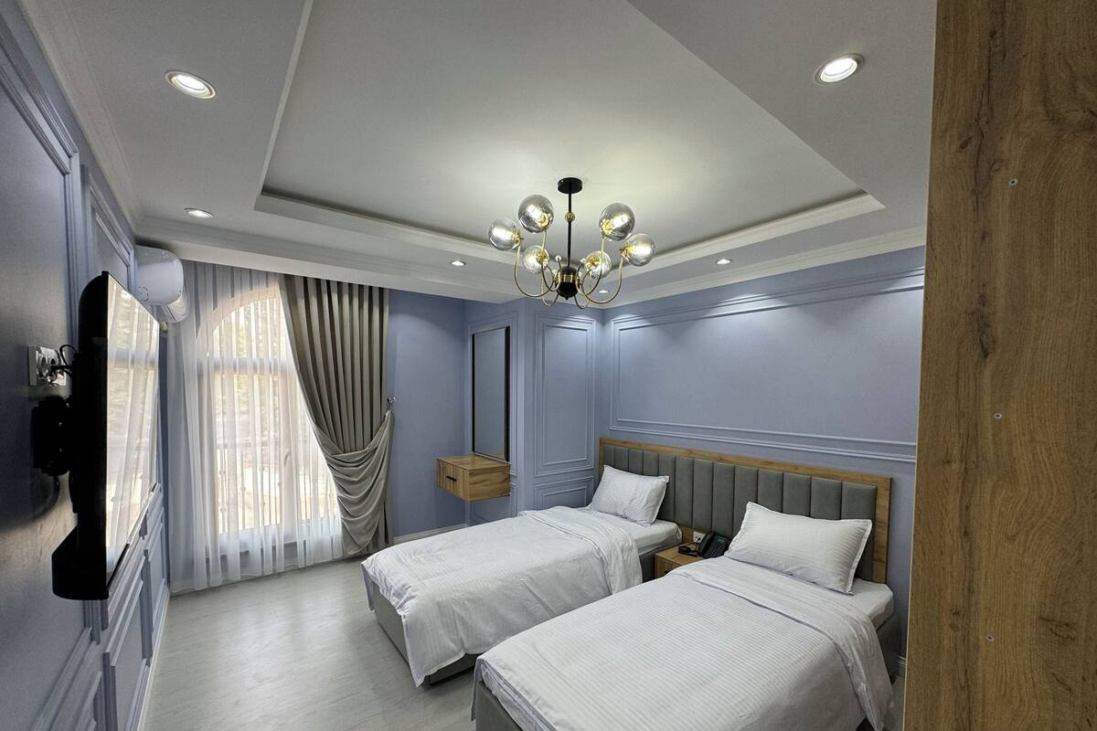 A hotel room with two beds, a vanity with a mirror, a chandelier, and a window with curtains.