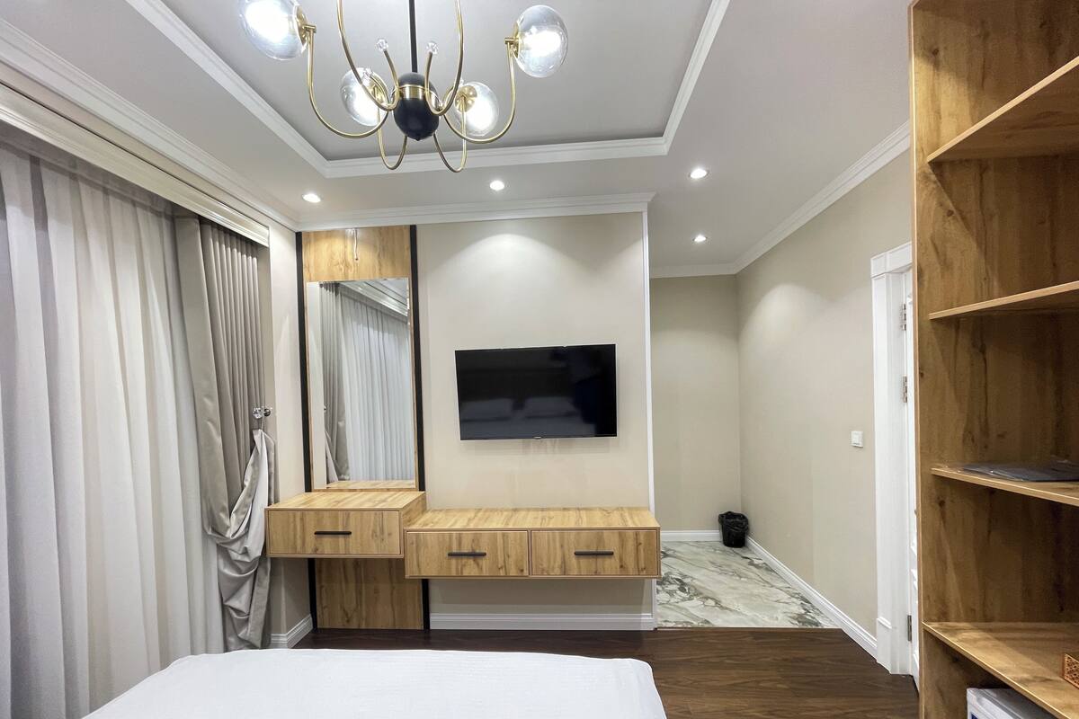 A bedroom with a bed, a TV mounted on the wall, a wooden vanity, and a chandelier.