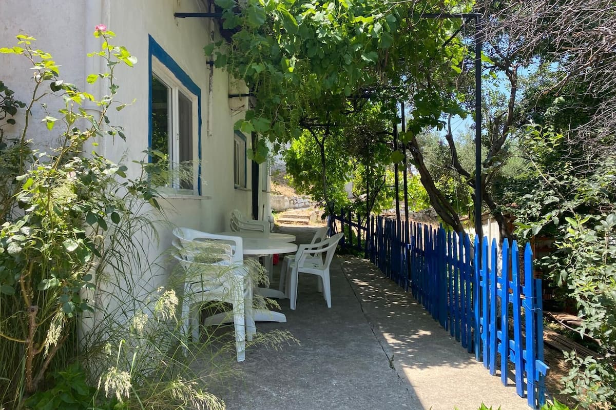 A covered outdoor seating area with white plastic chairs and a table, surrounded by a blue fence and greenery.