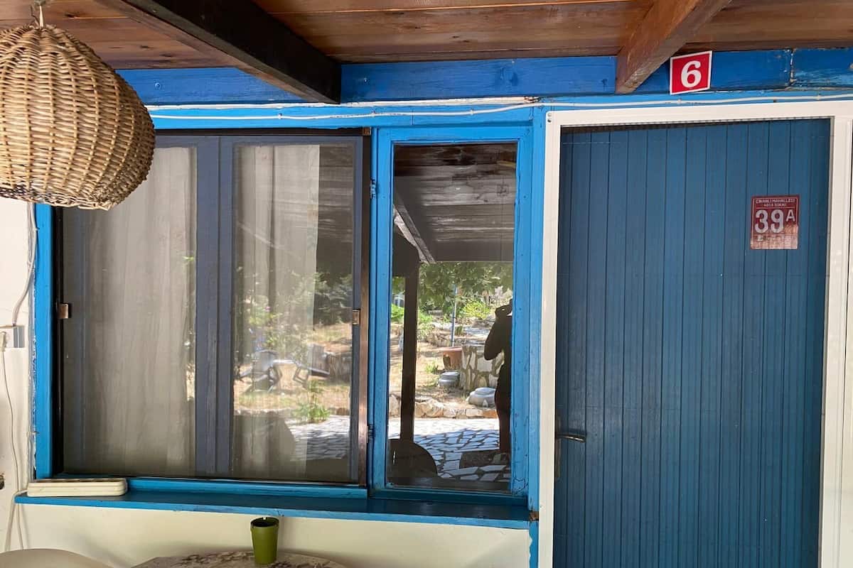 A room with a table, a chair, a wicker hanging light fixture, a blue door with a number sign, and a window with a view of the outside.