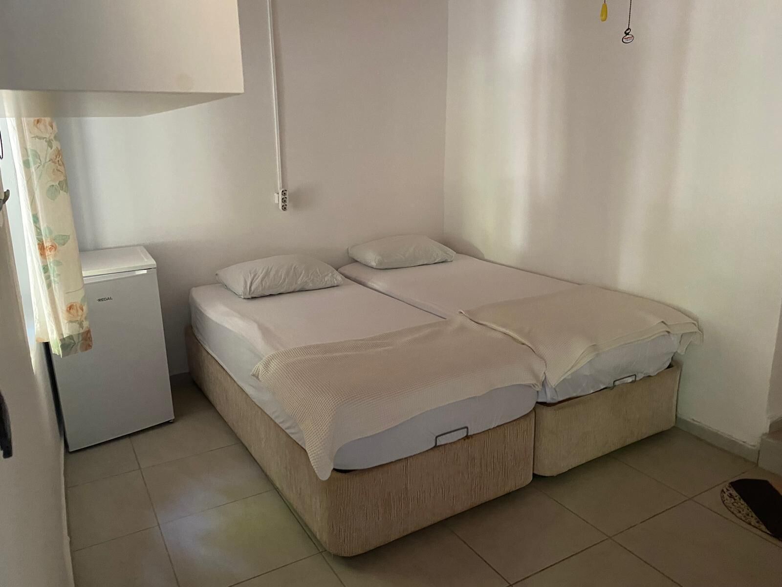 A single-bed room with a small refrigerator, a bedside table, and a wall-mounted light fixture.