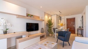 En moderne stue med et fladskærms-tv, træhylder, en stor stueplante, et spiseområde med et bord og stole samt et hyggeligt siddeområde med en sofa og lænestole.