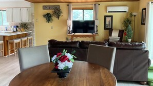 A cozy living room with a wooden ceiling, a TV on a wooden stand, a sofa, and a dining table with a floral centerpiece. - Cozy 3bedroom home w AC, WiFi, in a golf retreat, nestled in the scenic mountain (Basye)