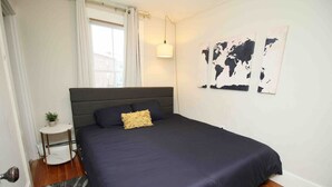 A bedroom with a dark blue bedspread, a small round side table with a plant, and a world map artwork on the wall.