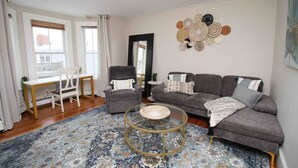 A living room with a grey sofa, a round glass coffee table, a white desk, and a chair.