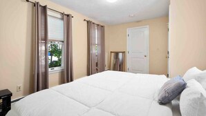 Un dormitorio con una cama grande, paredes color beige, una ventana con cortinas y una puerta.