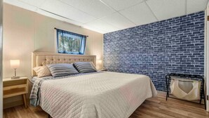 Een slaapkamer met een bed, nachtkastje, lamp, raam met blauw gordijn, een muur met baksteenmotief en een houten vloer.
