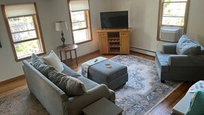A living room with a sofa, ottoman, and two armchairs, a TV on a wooden stand, and a window with blinds.