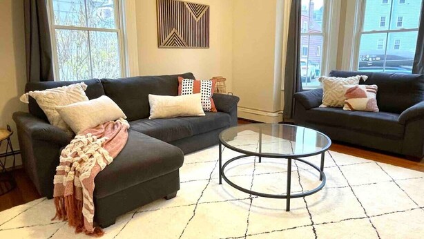 A living room with two dark-colored sofas, a glass coffee table, and a patterned rug. - 2 BR City Sanctuary - 5 Mins to Historical Salem! (Beverly)