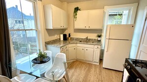 A compact kitchen with white cabinets, a gas stove, a refrigerator, and a dining area with a glass table and chairs. - 2 BR City Sanctuary - 5 Mins to Historical Salem! (Beverly)