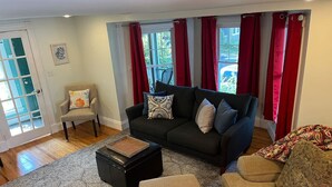 A living room with a dark-colored sofa, a beige armchair, and a patterned rug. There is a black ottoman in front of the sofa and a window with red curtains. - Swampscott! Walk Dtwn and beach (Swampscott)