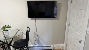 A room with a flat-screen TV mounted on the wall, a glass table with a chair, and a white door. - Dwtn Apt~King Beds~Mins to Salem (Beverly)