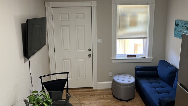 A room with a white door, a window with blinds, a blue couch, a small round ottoman, a television mounted on the wall, and a potted plant. - Dwtn Apt~King Beds~Mins to Salem (Beverly)
