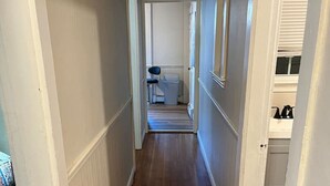 Een gang met houten vloeren en witte wanden, die leidt naar een kamer met een bed en een stoel.