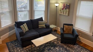 A living room with a dark-colored sofa, a matching armchair, a small coffee table, and a patterned rug. There are three windows with blinds, a floor lamp, and a painting on the wall. - Beverly/Salem! Dwtn/Water/Train (Beverly)