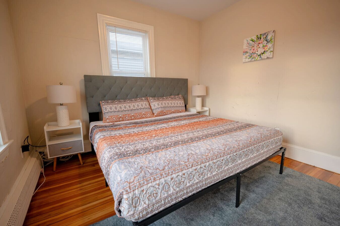 Une chambre à coucher avec une tête de lit capitonnée, une couvre-lit à motifs, deux lampes de chevet, une petite table de chevet blanche, une fenêtre avec des stores et une œuvre d’art florale accrochée au mur.