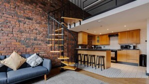 A modern loft with exposed brick walls, a staircase, a kitchen with wooden cabinets, and a blue sofa with cushions. - Liverpool Warehouse Flat | Great Location | Sleeps 5 | WiFi & Free Parking (Liverpool)