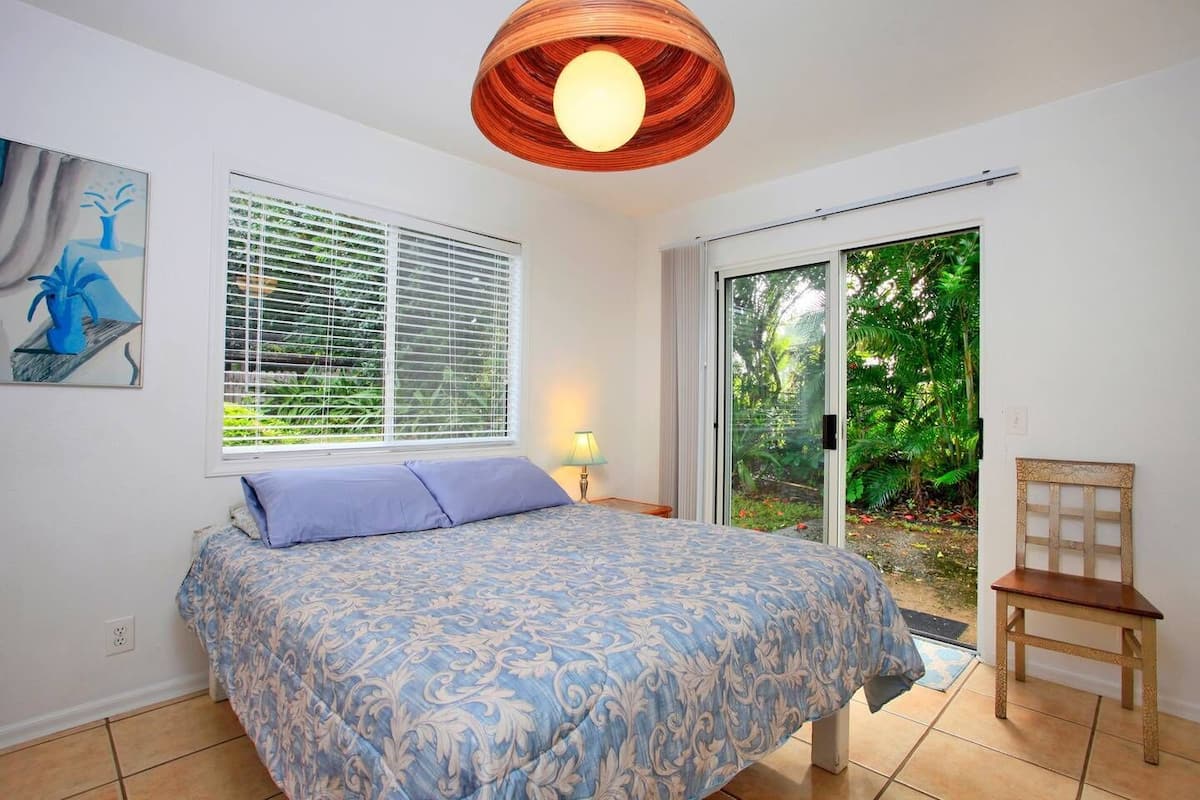 A bedroom with a bed, a window with blinds, a door leading to a garden, and a wooden chair.