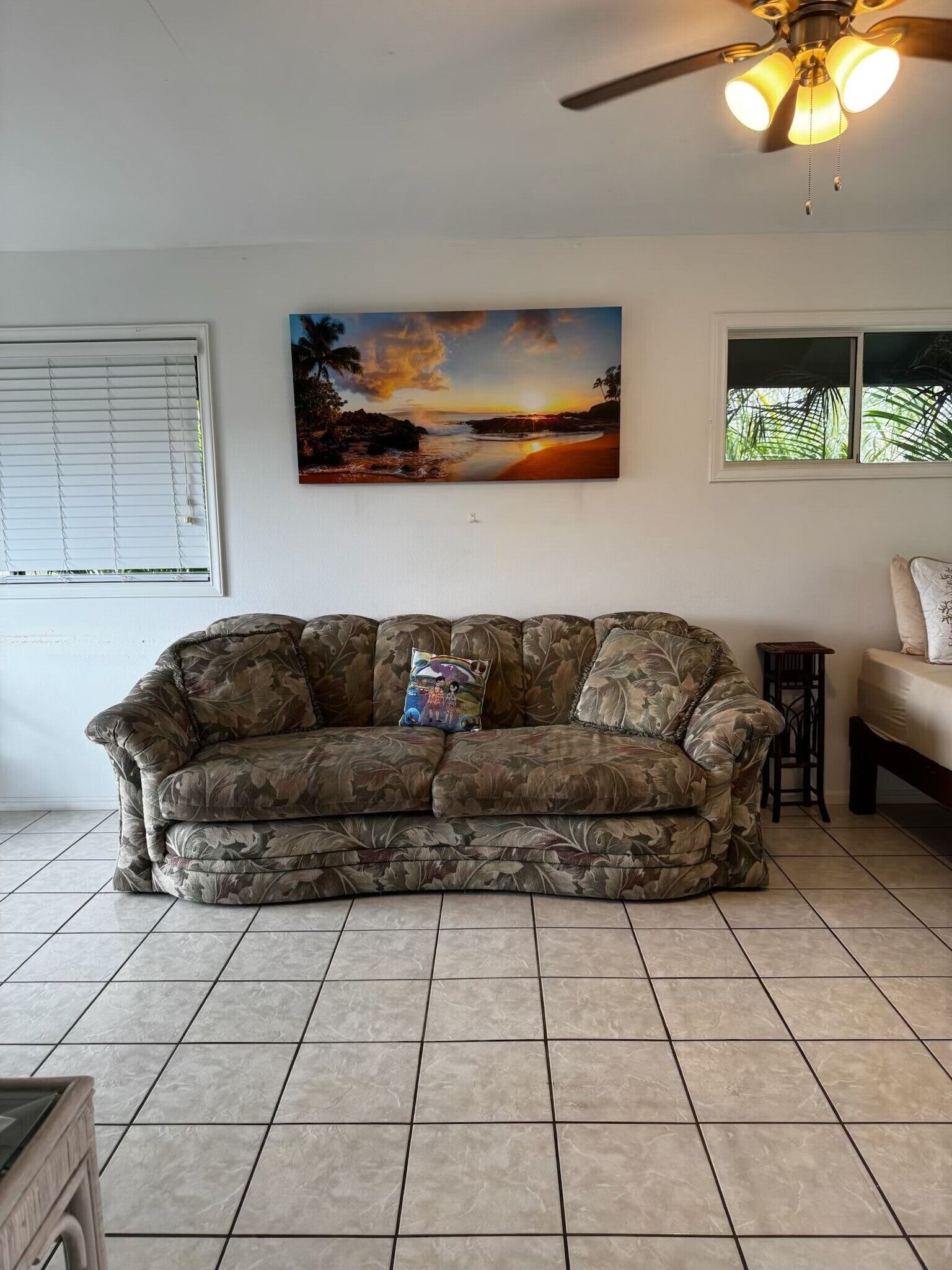 A living room with a couch, a ceiling fan, a painting of a sunset, and a window with blinds.