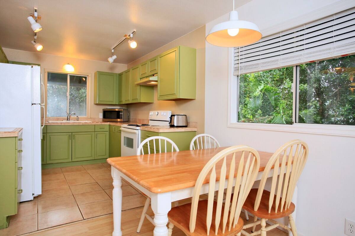 A compact kitchen with green cabinets, a wooden dining table, and white chairs. There is a window with blinds, a refrigerator, and a stove.