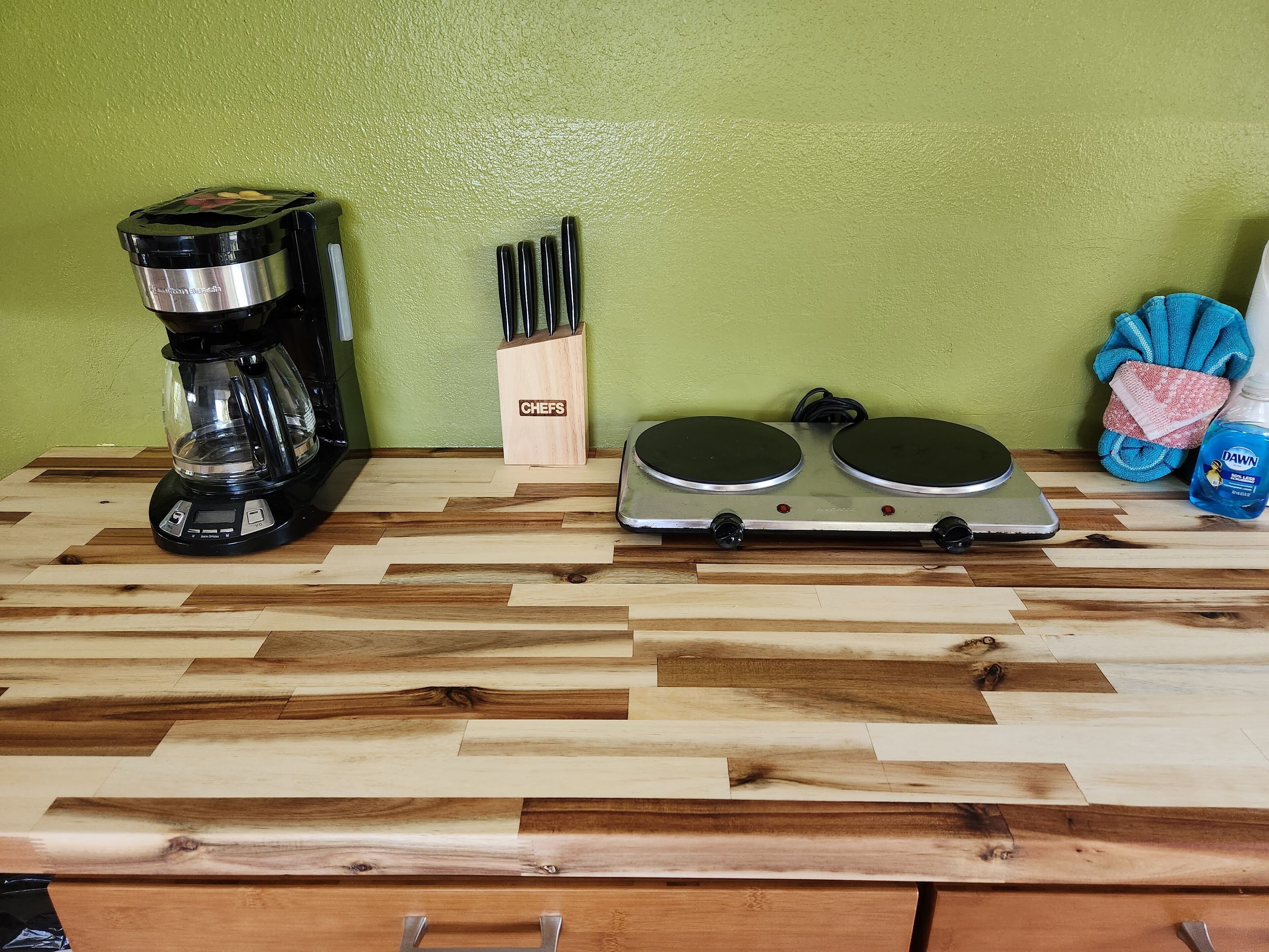 A kitchen counter with a coffee maker, knife block, electric griddle, and cleaning supplies.