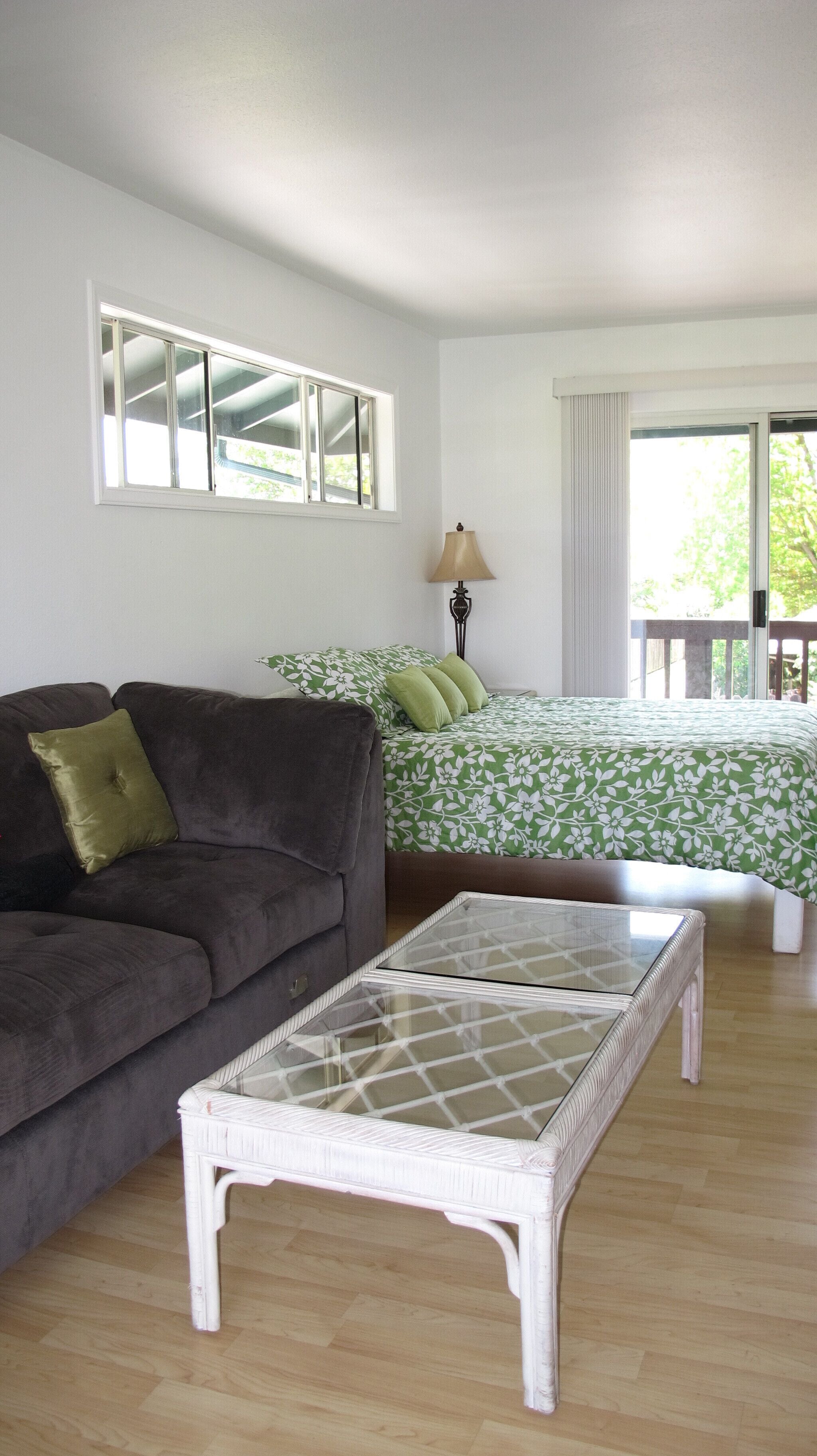 A room with a brown sofa, a white coffee table, a bed with green floral bedding, and a window with curtains.