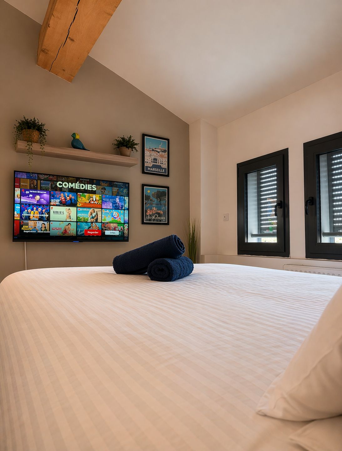 Une chambre d’hôtel avec un lit, un téléviseur fixé au mur diffusant une chaîne de comédies, une fenêtre avec des stores et un plafond avec une poutre en bois.