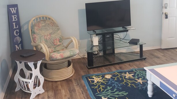 Een woonkamer met een televisie op een glazen standaard, een armstoel met bloemenprint, een klein wit bijzettafeltje, een welkomstbord en een blauw vloerkleed.