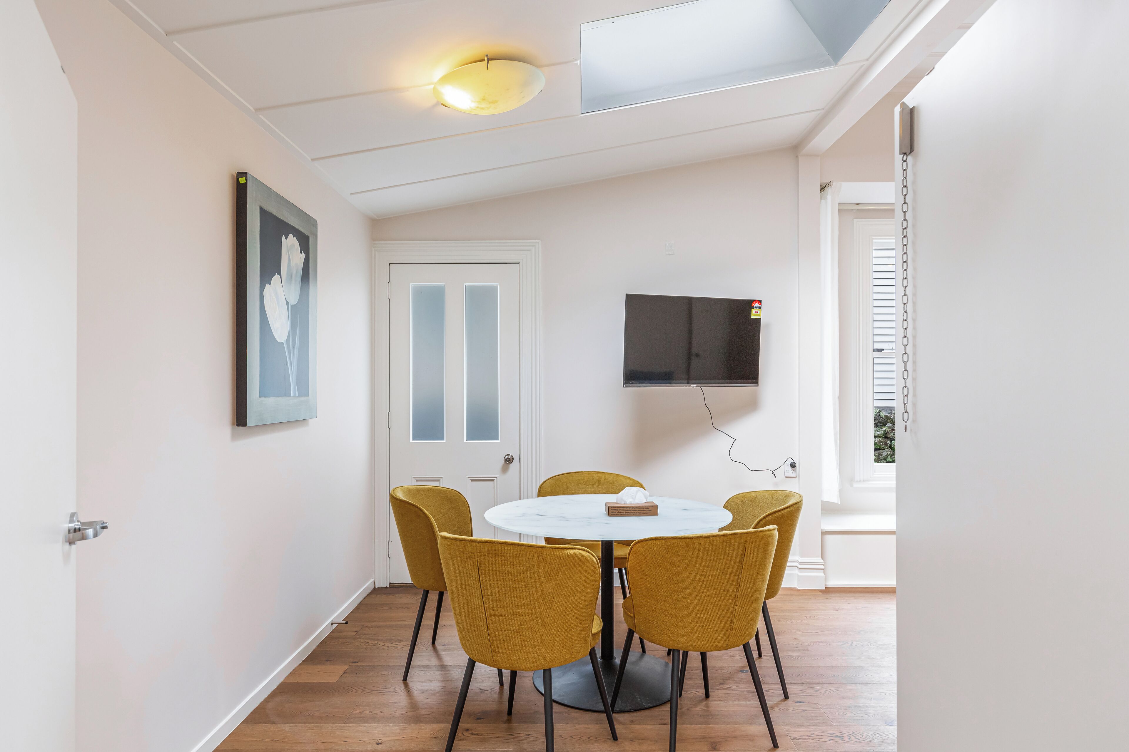 A dining area with a round table, six yellow chairs, a white door, and a mounted TV.