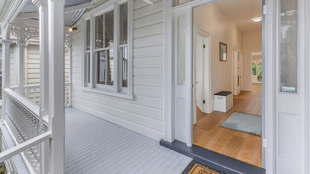 Une véranda en bois peinte en blanc, dotée d’une zone abritée, une porte donnant sur un couloir au plancher en bois, et une petite poubelle placée près de la porte.