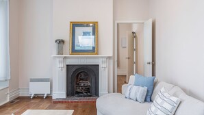 Une pièce avec une cheminée, un tableau encadré au-dessus, un radiateur blanc et un canapé avec des coussins.