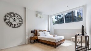 Een moderne slaapkamer met een grote wandklok, een bed met houten hoofdeinde, een nachtkastje met een lamp en een raam met uitzicht op gebouwen.