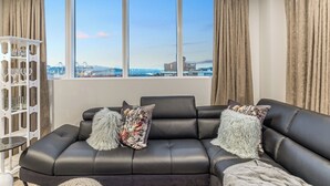 A modern living room with a black leather sofa, two decorative pillows, a glass coffee table, and a white side table. A large window offers a view of a cityscape and a bridge. - Modern 2BR Penthouse for Families by Zodiak Stays (Auckland)