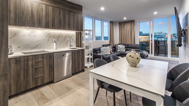 A modern kitchen with dark wood cabinets, a marble countertop, and stainless steel appliances. There is a dining area with a white table and chairs, and a living room with a black sofa and a large window offering a city view. - Modern 2BR Penthouse for Families by Zodiak Stays (Auckland)