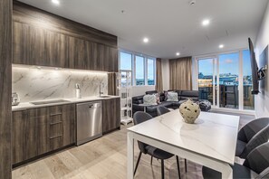 A modern kitchen with dark wood cabinets, a marble countertop, and stainless steel appliances. There is a dining area with a white table and chairs, and a living room with a black sofa and a large window offering a city view. - Modern 2BR Penthouse for Families by Zodiak Stays (Auckland)