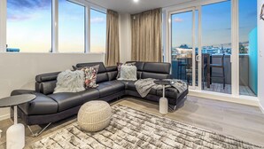A modern living room with a black leather sectional sofa, a round coffee table, and a large window offering a city view. - Modern 2BR Penthouse for Families by Zodiak Stays (Auckland)