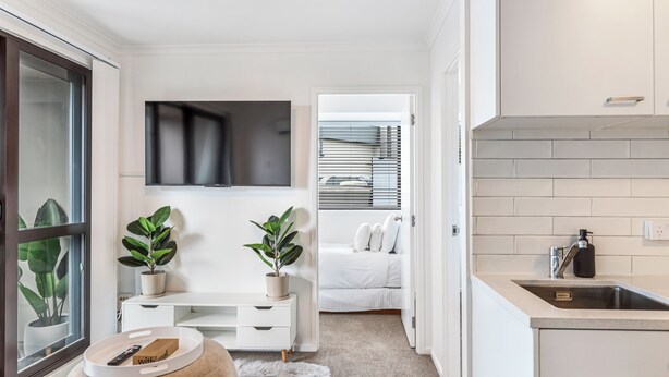 A modern, well-lit room with a white color scheme, a flat-screen TV mounted above a white console table, a small round ottoman with a remote control, and a potted plant. - Central 3BR Full Kitchen & Amenities Zodiak Stays (Auckland)