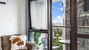 A balcony with a view of a cityscape, a potted plant, a wicker chair, and a brown sofa with a decorative pillow. - Central 3BR Full Kitchen & Amenities Zodiak Stays (Auckland)