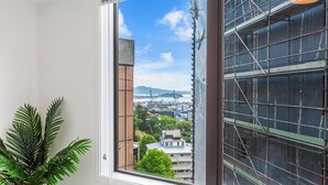 A window with a view of a cityscape, including buildings and a crane, and a potted plant to the left. - Central 3BR Full Kitchen & Amenities Zodiak Stays (Auckland)