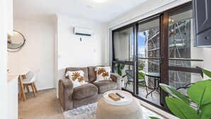 A modern living room with a brown sofa, a white coffee table, and a white rug. There is a dining area with a white table and chairs, and a large window with a view of construction outside. - Central 3BR Full Kitchen & Amenities Zodiak Stays (Auckland)