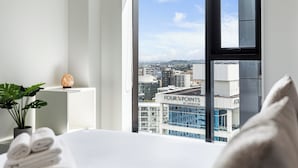 A hotel room with a large window offering a city view, a bed, a plant, and a decorative item on the windowsill. - Pool, Gym & Modern 2BR Sky Tower View Zodiak Stays (Auckland)