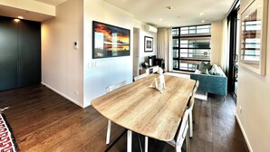 A modern living room with a wooden dining table, a sofa, and large windows offering a view. - 1BR with Balcony Close to Ferry by Zodiak Stays (Auckland)