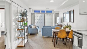 A modern living room with a sofa, a dining table with chairs, a kitchen area, and a large window with curtains. - 2BR Apt with Parking in Prime Spot Zodiak Stays (Auckland)