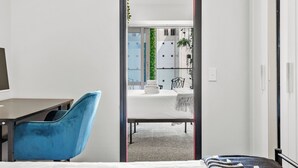 A modern hotel room with a bed, a desk, and a blue chair. There is a view of a dining area with a table and chairs. - 2BR Apt with Parking in Prime Spot Zodiak Stays (Auckland)