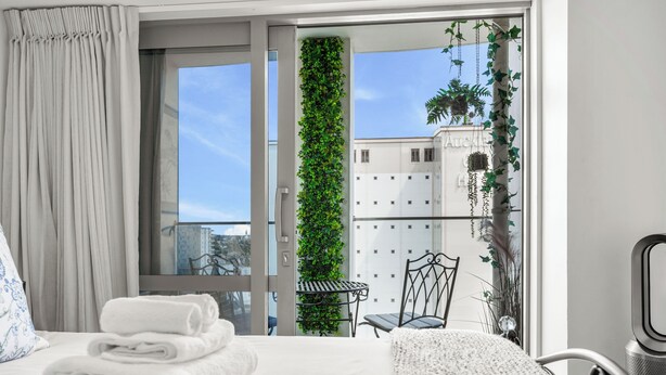 A hotel room with a bed, a neatly folded stack of towels, a view of the outside balcony with plants, and a chair. - 2BR Apt with Parking in Prime Spot Zodiak Stays (Auckland)
