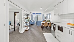 A modern kitchen with white cabinetry, a dining area with a table and chairs, and a living room with a blue sofa and a bookshelf. - 2BR Apt with Parking in Prime Spot Zodiak Stays (Auckland)