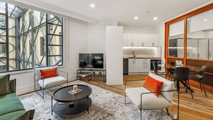 A modern living room with a sofa, coffee table, and TV, connected to a kitchen with white cabinets and a dining area. - Stunning 1BR Apt in Heritage Hotel by Zodiak Stays (Auckland)