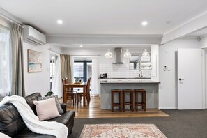 A modern living room with a leather sofa, a dining area with wooden chairs, a kitchen with white cabinets and pendant lights, and a hallway leading to a door.