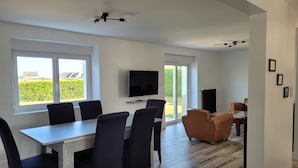 Ein modernes Wohnzimmer mit Essbereich, großen Fenstern, einem Flachbildfernseher und einem Kamin.
